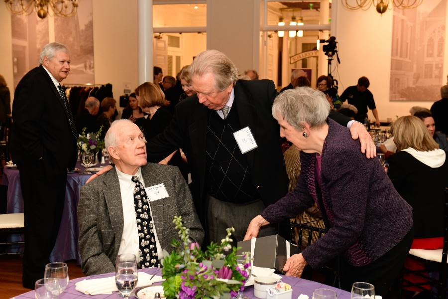 Three people welcome each other at a table with a purple tablecloth covering it.